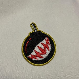 May include: Embroidered patch featuring a black bomb design with a red and white grinning mouth. The patch has a yellow border and a twisted rope fuse. This patch is suitable for jackets, bags, or other fabric items.