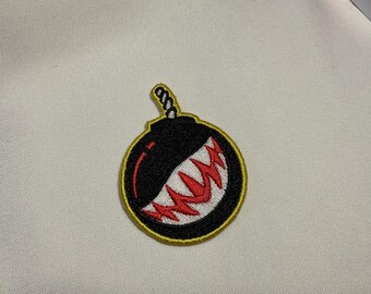 May include: Embroidered patch featuring a black bomb design with a red and white grinning mouth. The patch has a yellow border and a twisted rope fuse. This patch is suitable for jackets, bags, or other fabric items.