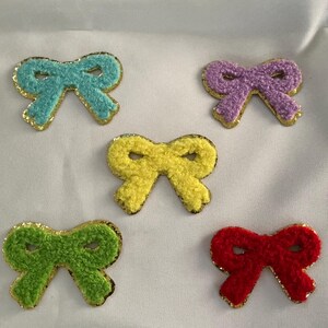 May include: Five colorful bow-shaped patches. The bows are in shades of blue, purple, yellow, green, and red. Each bow has a textured, fuzzy appearance and is outlined with gold trim. These patches are suitable for various craft projects.