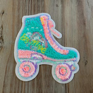 May include: A colorful, sequined roller skate patch. The skate features a teal upper, pink accents, and purple and pink wheels. The design is outlined in white, creating a playful and vibrant aesthetic.