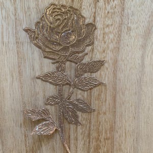 May include: A detailed, metallic rose sculpture in a gold color. The rose has intricate petal and leaf detailing, with a stem and leaves. The sculpture is placed on a wooden surface.