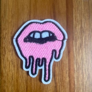 May include: Pink lips with a black outline and dripping effect. The lips are embroidered on a white fabric patch.
