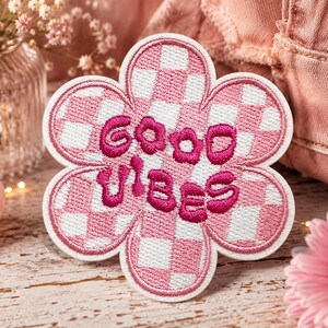 May include: A flower-shaped embroidered patch featuring a pink and white checkered pattern. The center of the patch displays the text "good vibes" stitched in a bold, dark pink thread with a rounded, bubbly font style and a white border.