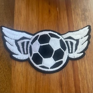 May include: A black and white embroidered patch of a soccer ball with wings. The soccer ball is in the center of the patch and the wings are on either side of it.