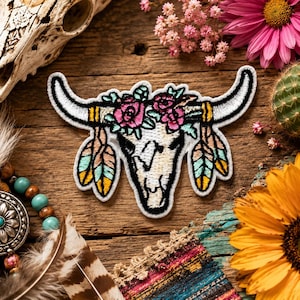 May include: An embroidered patch featuring a white bull skull with long curved horns, adorned with pink roses and green leaves. The design includes dangling feathers in shades of yellow, pink, and teal, outlined in black thread on a white fabric base.