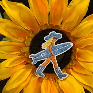 May include: Embroidered patch featuring a surfer girl in a cowboy hat and boots, carrying a surfboard. The patch is blue, orange, and white, set against a bright yellow sunflower with a dark brown center. The patch is likely for clothing or accessories.