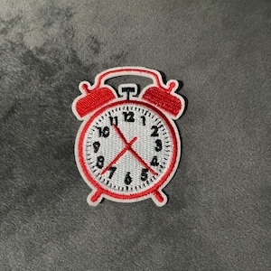May include: Embroidered patch in the shape of a red alarm clock. The clock face is white with black numbers and hands, and the clock's outline is red. The patch is designed to be ironed or sewn onto fabric.