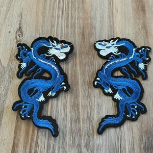 May include: Two embroidered patches featuring a blue dragon design with white accents. The dragon is depicted in a curved pose with intricate details, set against a black background. The patches are placed on a wooden surface.
