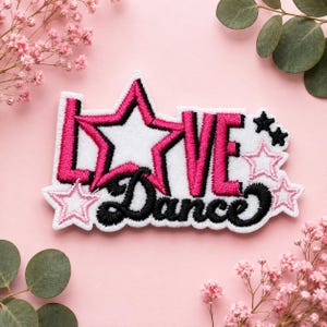 May include: An embroidered patch featuring the word LOVE with a large pink and black star replacing the letter O, and the word Dance in black cursive script below. The design includes additional small pink and black stars on a white background.