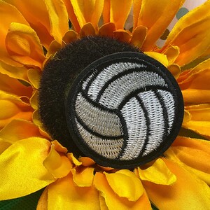 May include: A circular embroidered patch featuring a white and silver volleyball design with black stitching details and a solid black border, centered on a large yellow sunflower with prominent petals and a dark brown textured center.