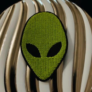 May include: Green alien face embroidered patch with black eyes. The patch is on a white and gold striped background.