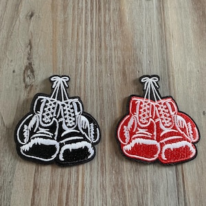 May include: Two embroidered patches shaped like pairs of boxing gloves. One patch features black gloves with white laces and trim, while the other patch features red gloves with white laces and trim. Both patches have a black border outline.