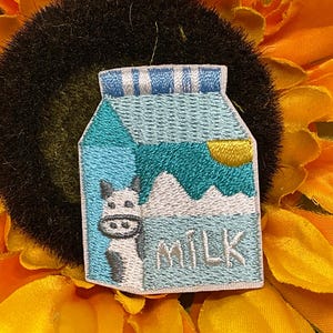 May include: An embroidered patch shaped like a milk carton featuring a light blue and teal color scheme. The design includes a white cow with black spots, a yellow sun, white mountain peaks, and the word MILK stitched in white thread.