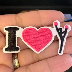 May include: Embroidered patch with the text "I ♥ NY" in black, pink, and white. The "I" is black, the heart is pink, and the "NY" is a black stick figure with pink accents. The patch has a white border.