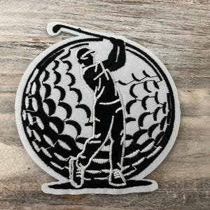 May include: Black and white embroidered patch featuring a golfer mid-swing. The design is set within a golf ball shape, with the golfer's silhouette and golf club clearly defined. The patch is likely for apparel or accessories.