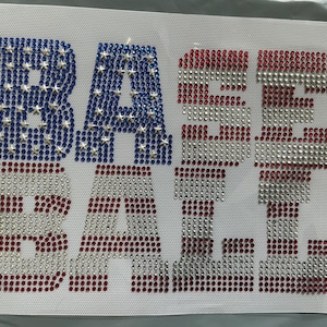 May include: A rhinestone iron-on transfer featuring the word BASEBALL in block letters. The letters B, A, and S are filled with blue rhinestones and silver star accents, while the remaining letters feature red and white horizontal stripes.