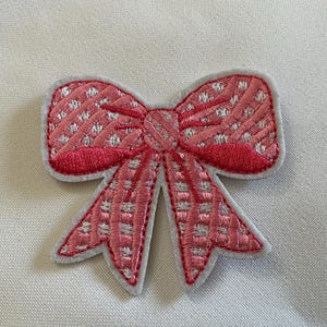 May include: A pink embroidered bow patch with a white border. The bow features a textured pattern with white accents. The bow's design includes a central knot and two loops on each side, with two tails extending downwards.
