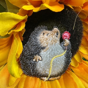 May include: Embroidered hedgehog patch holding a red flower, set against a bright yellow sunflower. The hedgehog has gray and brown fur with a light tan belly. The sunflower petals are a vibrant yellow.