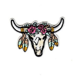 May include: Embroidered patch of a white cow skull with pink flowers and turquoise and yellow feathers.