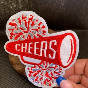 May include: A red and white embroidered patch featuring a megaphone with the word "CHEERS" in white lettering. The patch includes two pom-poms with red and silver accents, perfect for cheerleading or spirit wear.