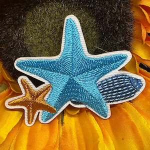May include: Embroidered patches featuring a large turquoise starfish, a small gold starfish, and a blue and white fish. The patches have white borders and are set against a yellow and green background.
