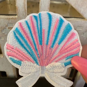 May include: Embroidered seashell patch with a white bow. The seashell design features pink and blue rays against a white background. This decorative patch is suitable for clothing, bags, or other craft projects.