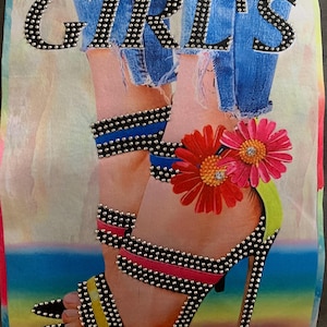 May include: A colorful graphic featuring the word "GIRLS" in silver studded letters. The image depicts a pair of high-heeled sandals adorned with studs and colorful straps, with a floral accent. The background shows a pair of blue jeans.