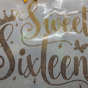 May include: Gold rhinestone design on a white background. The design features the words "Sweet Sixteen" in a script font, a crown, a butterfly, and decorative stars. The rhinestones are arranged to create the lettering and images.