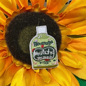 May include: An embroidered patch shaped like a bottle with a light green background and white cap. The text reads Beverage, Matcha, and Tea Drink. Small red and pink accents are scattered across the surface of the bottle-shaped patch design.