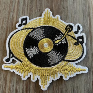 May include: Embroidered patch featuring a black and silver vinyl record with a gold needle and sound wave design. Musical notes and a white border complete the design. A music-themed patch.