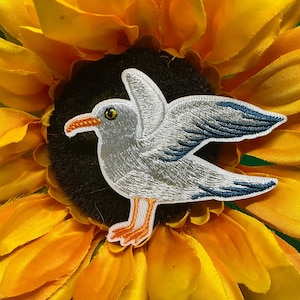 May include: Embroidered patch of a seagull with white, gray, blue, and orange details. The patch is set against a yellow and orange sunflower.
