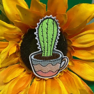 May include: Embroidered patch featuring a green cactus in a decorative pot. The patch has a white border and is set against a bright yellow sunflower. The pot is gray, brown, and pink.