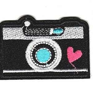 May include: Black and white embroidered iron-on patch of a camera with a blue lens and a pink heart.