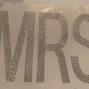 May include: The word "MRS" spelled out in silver rhinestones on a white background.