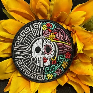May include: Round embroidered patch featuring a split design: a black and white Aztec-inspired pattern on one side and a colorful sugar skull on the other. The patch is set against a yellow sunflower.