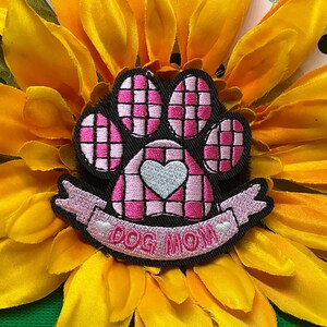 May include: An embroidered patch shaped like a paw print with a pink and white checkered pattern. A light gray heart is centered on the paw pad. Below the paw, a pink banner features the text DOG MOM in pink letters with two small hearts.