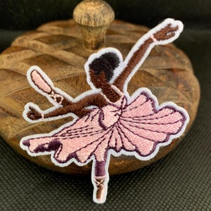 May include: Embroidered patch of a ballerina in a graceful pose. The patch features a pink tutu, brown arms, and a black bun. The ballerina is wearing ballet shoes. The patch is outlined in white and is on a wooden surface.