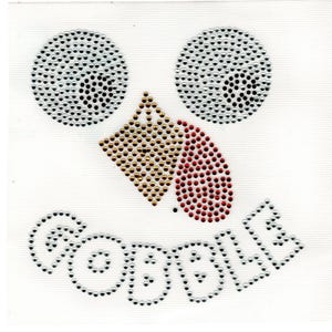 May include: A white fabric square featuring a face design made of rhinestones. The eyes are two circles of silver rhinestones, the nose is a gold diamond shape, and the beak is red. The word "GOBBLE" is spelled out in silver rhinestones.