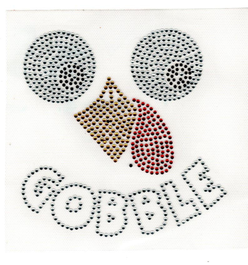 May include: A white fabric square featuring a face design made of rhinestones. The eyes are two circles of silver rhinestones, the nose is a gold diamond shape, and the beak is red. The word "GOBBLE" is spelled out in silver rhinestones.