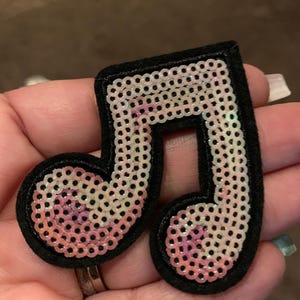 May include: A sequined musical note patch with a black border. The sequins are iridescent, with pink and white hues. The patch is a decorative item, likely for clothing or accessories.