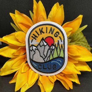 May include: An embroidered patch with the words "HIKING CLUB" in a yellow and blue oval design. The patch features a mountain range, a red sun, and green trees. The patch is set against a bright yellow sunflower.