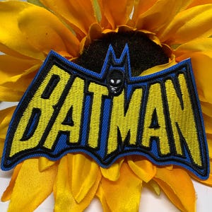 May include: Embroidered Batman patch with yellow lettering and a black outline on a blue background. The patch features a bat symbol and the word "BATMAN". The patch is set against a yellow flower.