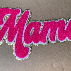 May include: A pink felt patch with the word "Mama" in a cursive font. The letters are outlined in white and edged with gold glitter. The patch is on a neutral background.