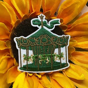 May include: An embroidered patch featuring a green carousel with gold detailing, white horses, and a small star topper. The design includes a green ribbon accent at the top, all rendered in detailed thread work on a white fabric backing.
