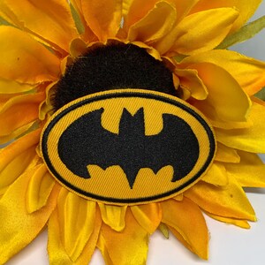 May include: A yellow and black Batman logo patch is centered on a bright yellow sunflower. The patch is oval-shaped with a black bat symbol on a yellow background. The sunflower has multiple layers of yellow petals.