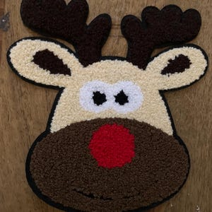 May include: A plush reindeer face with a red nose, brown antlers, and black eyes. The reindeer's face is beige with a brown muzzle and a black outline. This festive decoration is perfect for Christmas.