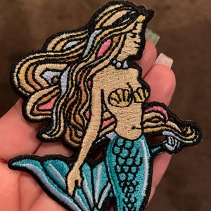 May include: An embroidered patch featuring a mermaid with long flowing blonde hair, a tan skin tone, and a seashell top. The mermaid has a bright turquoise tail with dark blue scale detailing and black outlines. The design is finished with a black border.