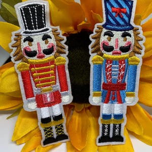 May include: Two embroidered nutcracker patches. One nutcracker wears a red uniform with a black hat, the other wears a blue uniform with a blue hat. Both have brown hair, black mustaches, and are set against a yellow sunflower.