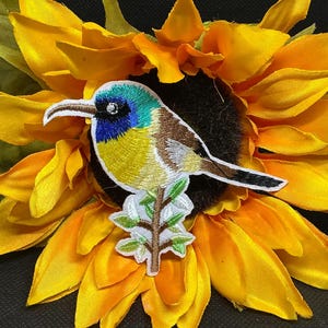 May include: Embroidered patch of a colorful bird perched on a branch, set against a bright yellow sunflower. The bird features blue, yellow, brown, and white details. The sunflower has vibrant yellow petals and a dark center.