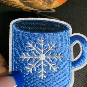 May include: Embroidered patch featuring a blue coffee mug with a white snowflake design. The mug has a white border and a blue handle. The patch is likely for clothing or accessories.